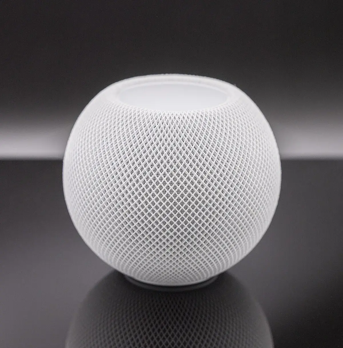 A white Apple HomePod mini smart speaker on a dark reflective background.