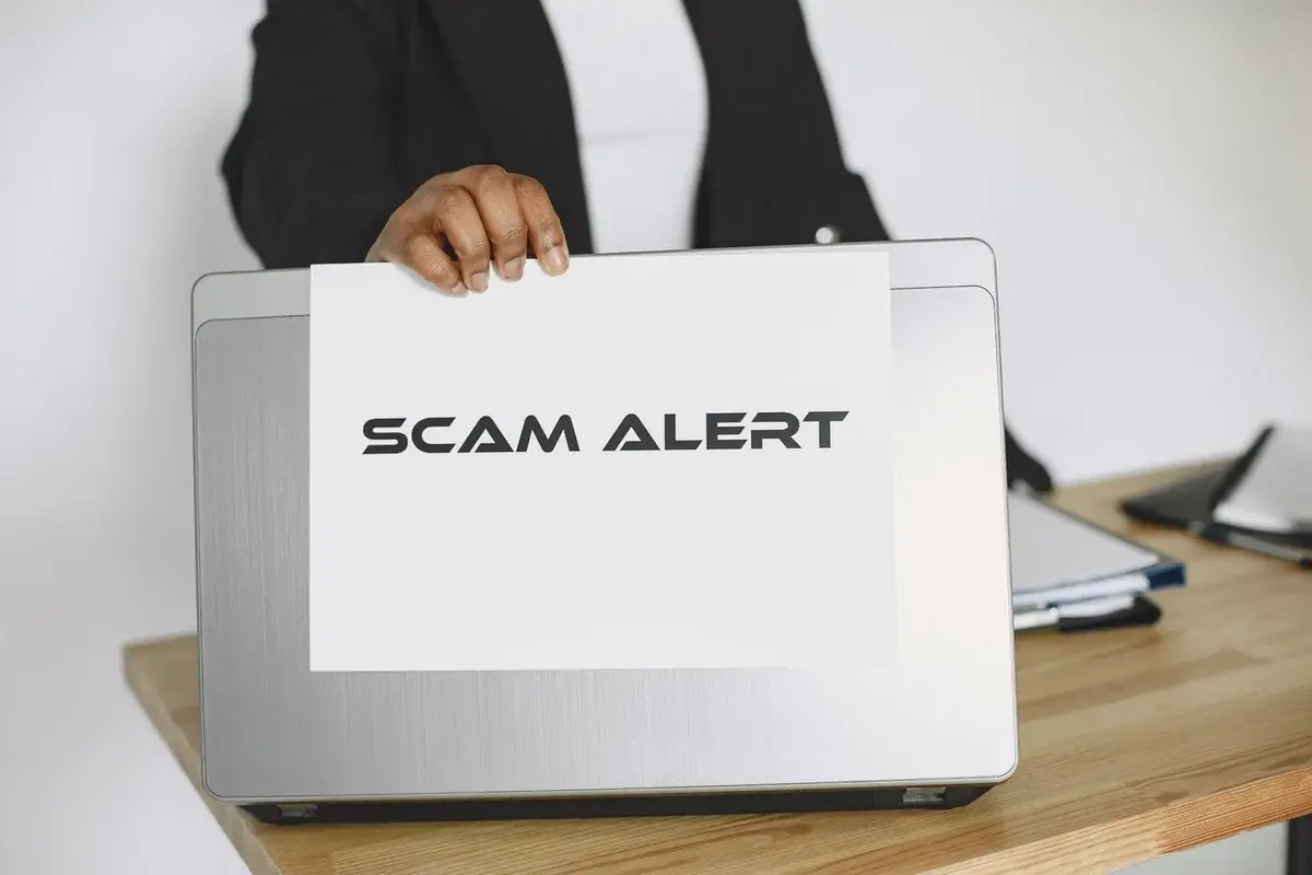 A person holds a sign that says "SCAM ALERT" in front of a laptop.