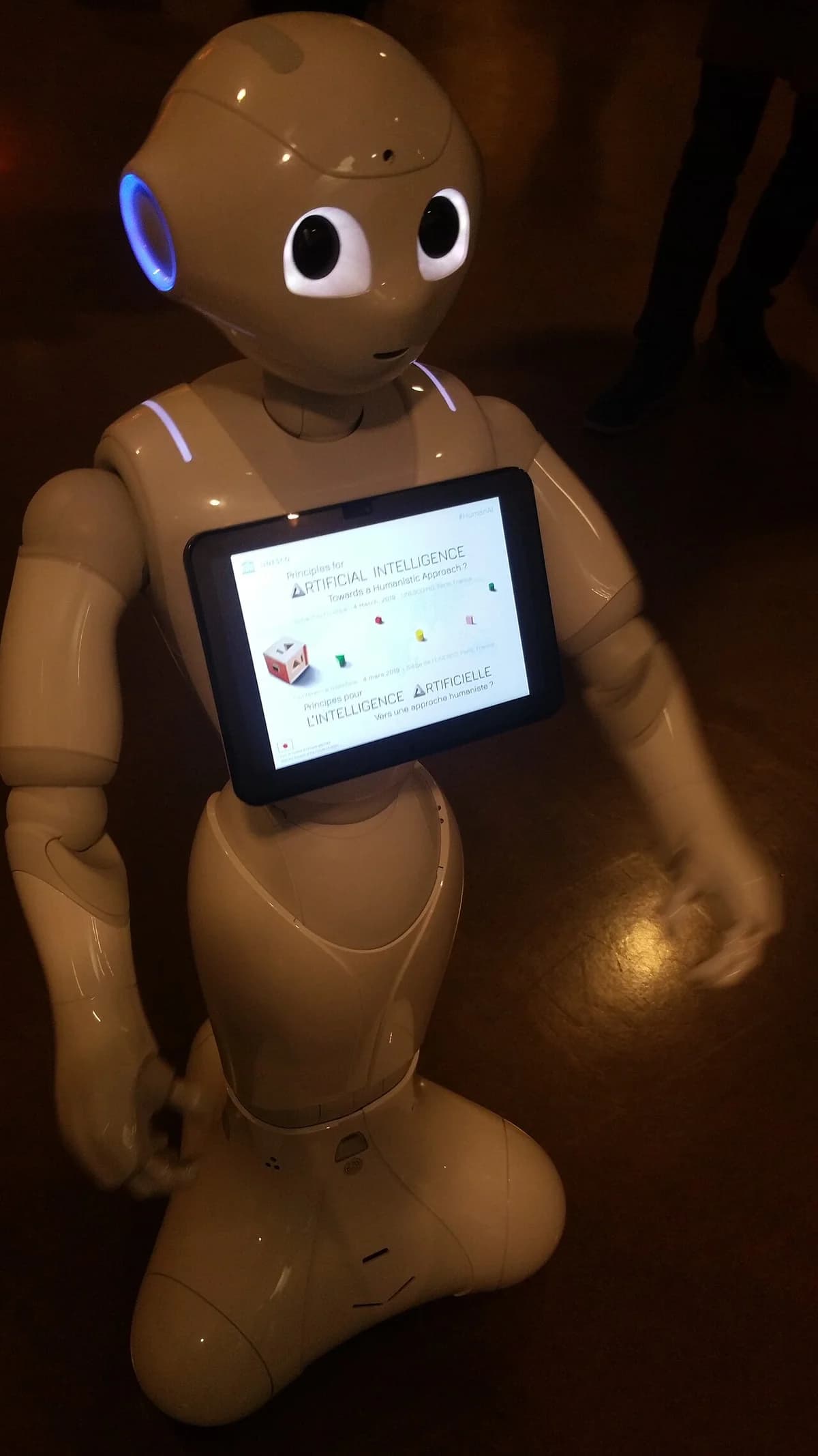 A humanoid robot in a conference setting with a tablet on its chest.