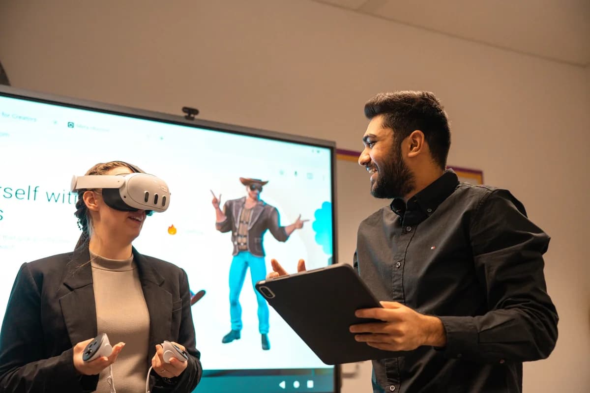 Two coworkers collaborating in an office, one using a VR headset and controllers while the other holds a tablet.