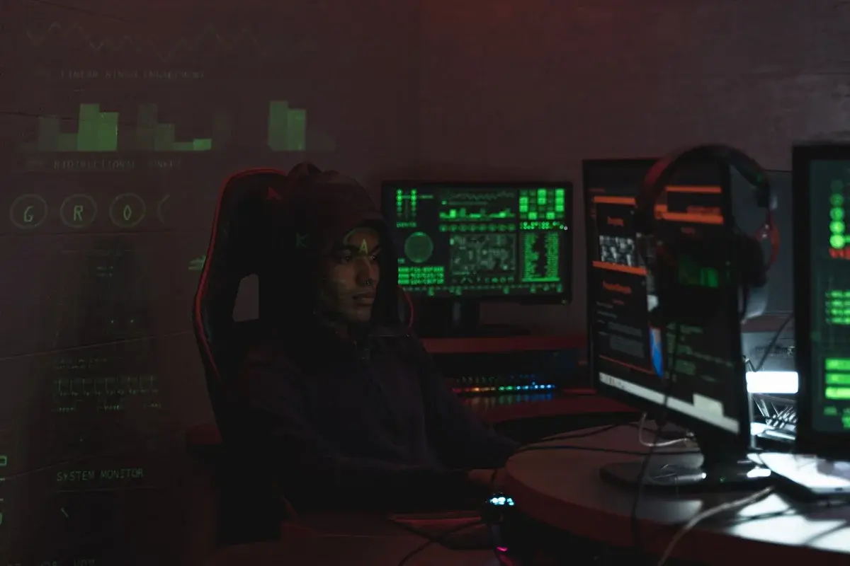 A Black man in a dark hoodie sits at a desk with multiple glowing computer monitors.
